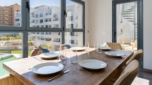 Een eetruimte met een houten tafel gedekt voor vier personen, grote ramen en uitzicht op een woongebouw en een zwembad.