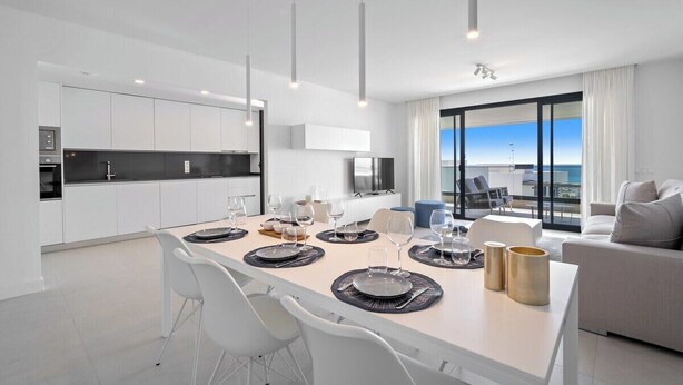 A modern kitchen with white cabinets, a dining area with a long table set for six, and a large window offering a view of the sea. - Apartment with sea view and gym (Estepona)
