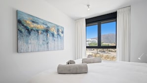 A modern bedroom with a large bed, a painting on the wall, and a view of mountains through the window. - Apartment with sea view and gym (Estepona)