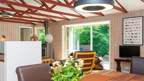 A modern interior with a wooden dining table, chairs, a vase with flowers, and a large window offering a view of greenery. - 4 Person Holiday Home in Glesborg (Glesborg)