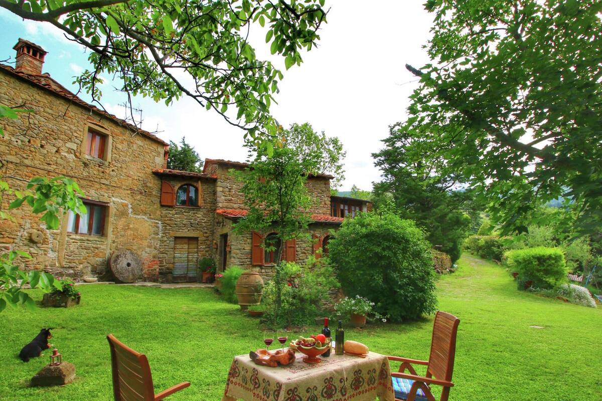 Ein Steinhaus mit einem Esstisch im Garten, umgeben von üppigem Grün und einem Hund, der auf dem Gras liegt.