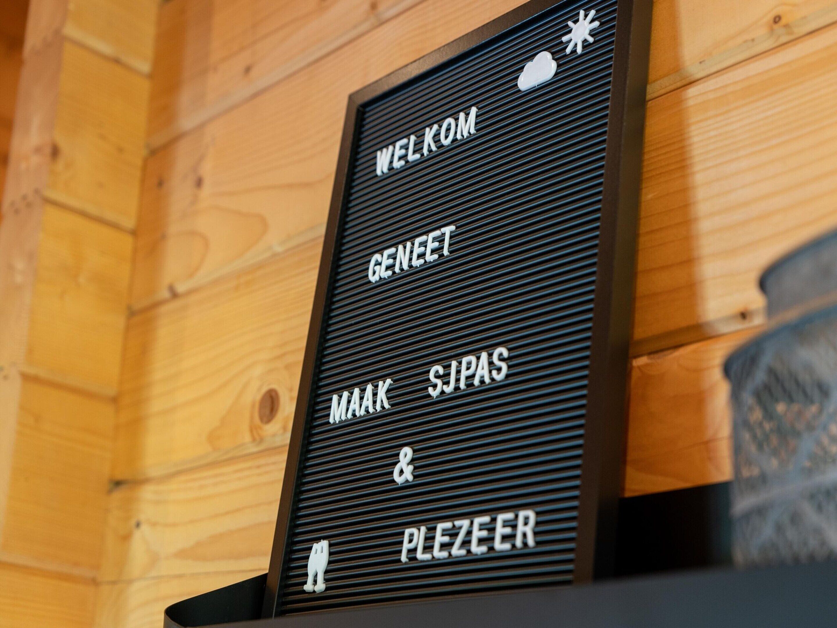 A welcome sign with the text "WELKOM GENET MAAK & SJPAS PLEZEER" in a wooden interior.
