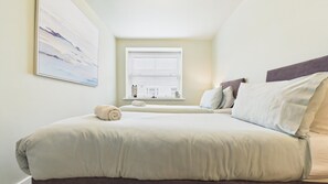 A bedroom with a large bed, a window with blinds, a painting on the wall, and a small table with a towel. - Dolphin - 2 Bedroom Apartment - Saundersfoot (Saundersfoot)