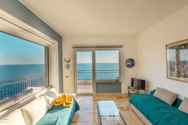 Une pièce dotée d’une grande fenêtre donnant sur la mer, d’un canapé recouvert d’un tissu turquoise, d’une table basse en verre et d’un téléviseur posé sur une table d’appoint.