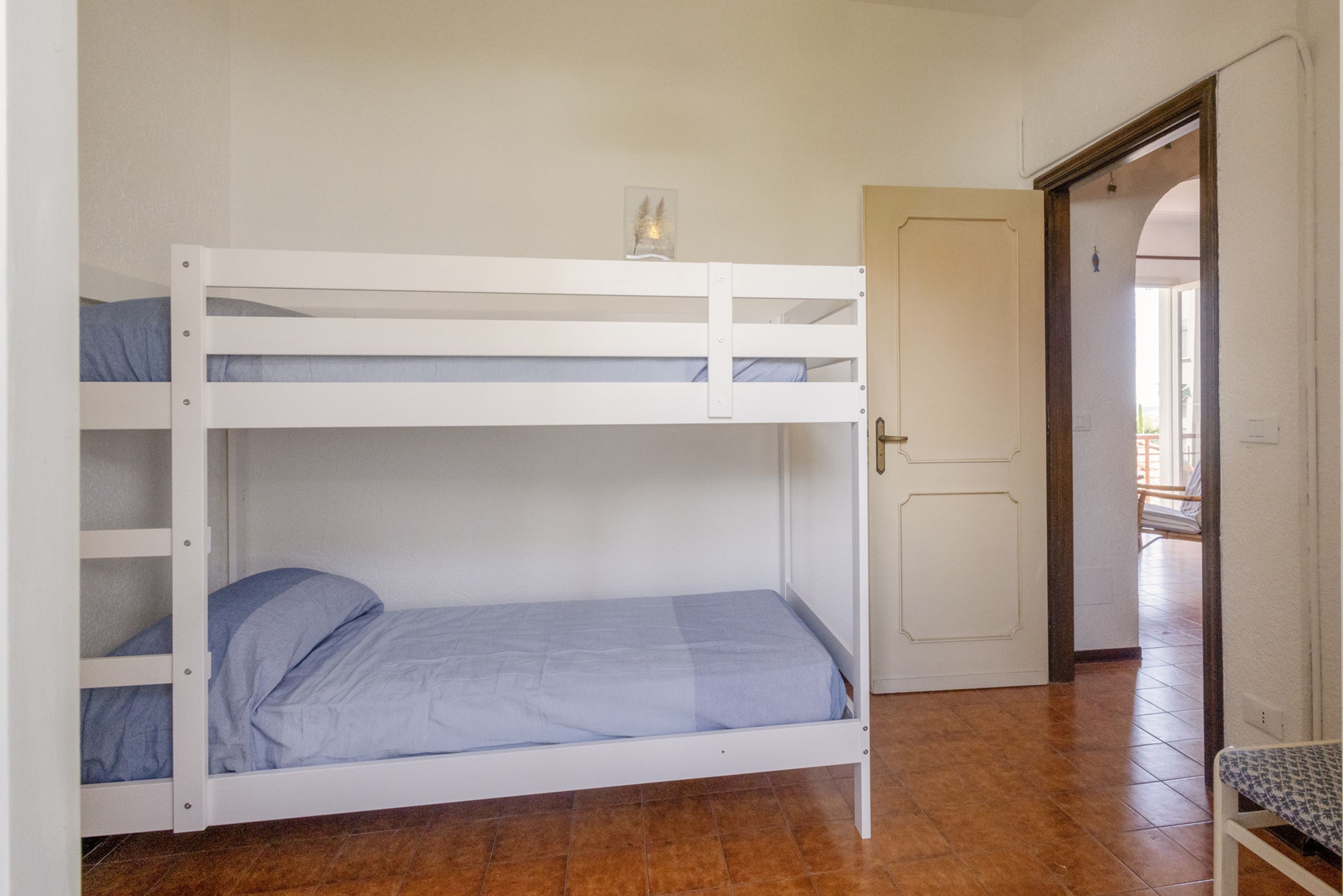 Un lit superposé avec un matelas bleu, un petit tableau encadré sur le lit du haut, et une porte donnant sur une autre pièce.