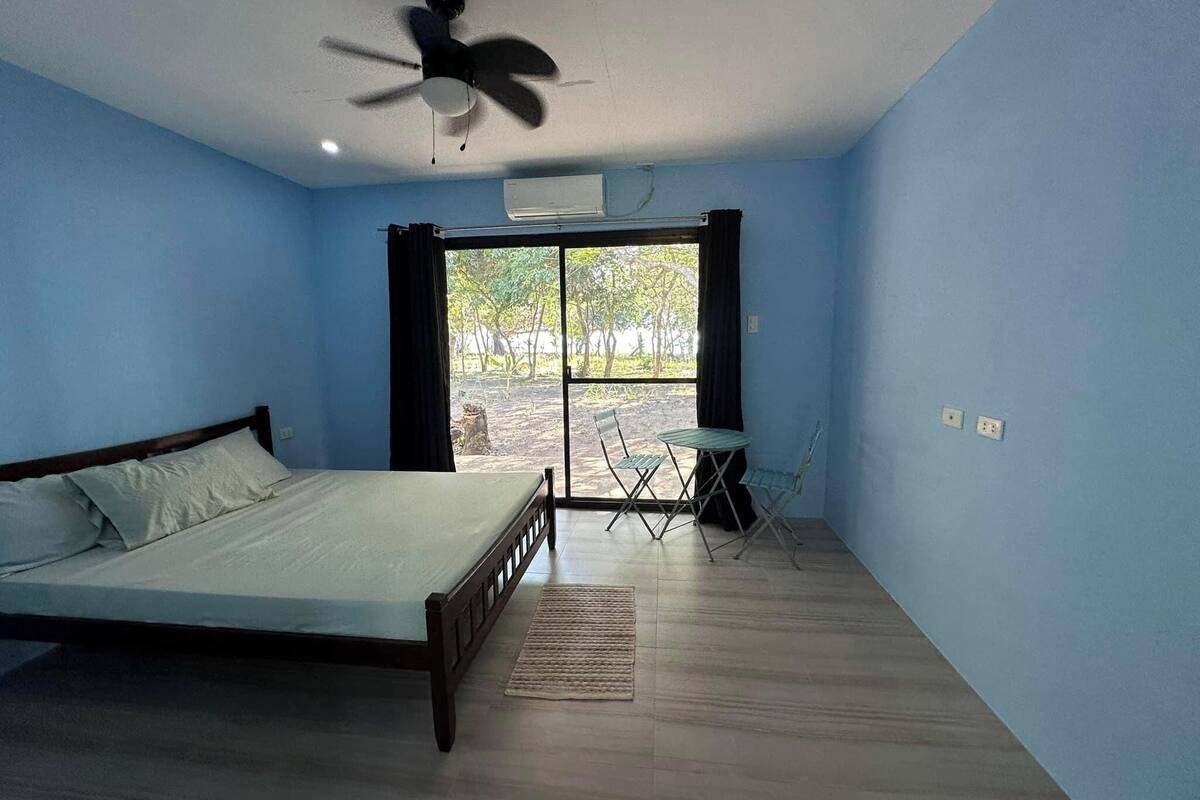 A room with a bed, a ceiling fan, a table with chairs, and a view through a glass door.