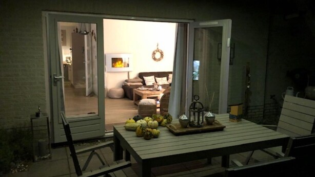 An outdoor patio area with a wooden table and chairs, a fire feature, and a view into the house. - Holiday Home Near Beach (Ouddorp)