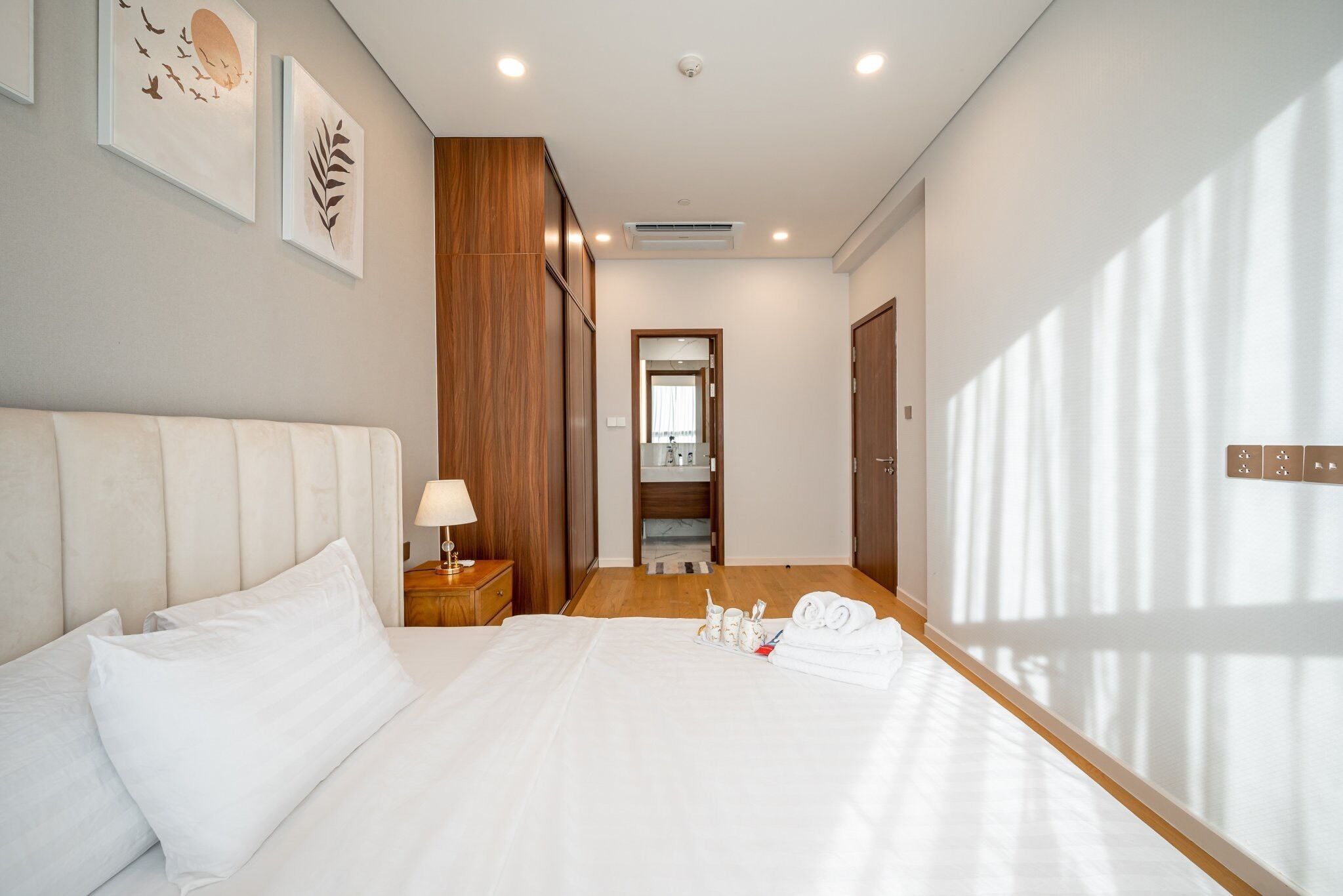 A hotel room with a large bed, a nightstand with a lamp, a wooden wardrobe, and framed artwork on the wall.