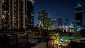 A nighttime view of a cityscape with modern buildings, a bridge, and a waterfront area with a playground. - Metropole Apartment Thu Thiem (Ho Chi Minh City)