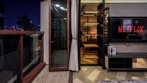 A modern living room with a large flat-screen TV displaying 'NETFLIX', a sliding glass door leading to an outdoor balcony, and a cityscape view at night. - Metropole Apartment Thu Thiem (Ho Chi Minh City)