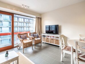 Ein modernes Wohnzimmer mit Fernseher, Korbsesseln, einem Esstisch und Blick auf ein Gebäude durch große Fenster.