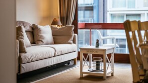 Un salon moderne avec un canapé, une petite table sur laquelle se trouve une lampe, et une vue sur le paysage urbain depuis un balcon.