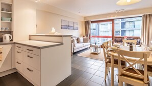 Un appartement moderne comprenant une cuisine, un coin repas et un salon. Il dispose d’une grande fenêtre offrant une vue sur les immeubles, d’un canapé et d’une table à manger avec des chaises.