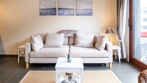 Un salon avec un canapé beige, une petite table blanche et une lampe. Trois tableaux sont accrochés au mur, au-dessus du canapé.
