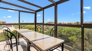 A sunlit room with a table and chairs, large windows offering a view of greenery and distant buildings.