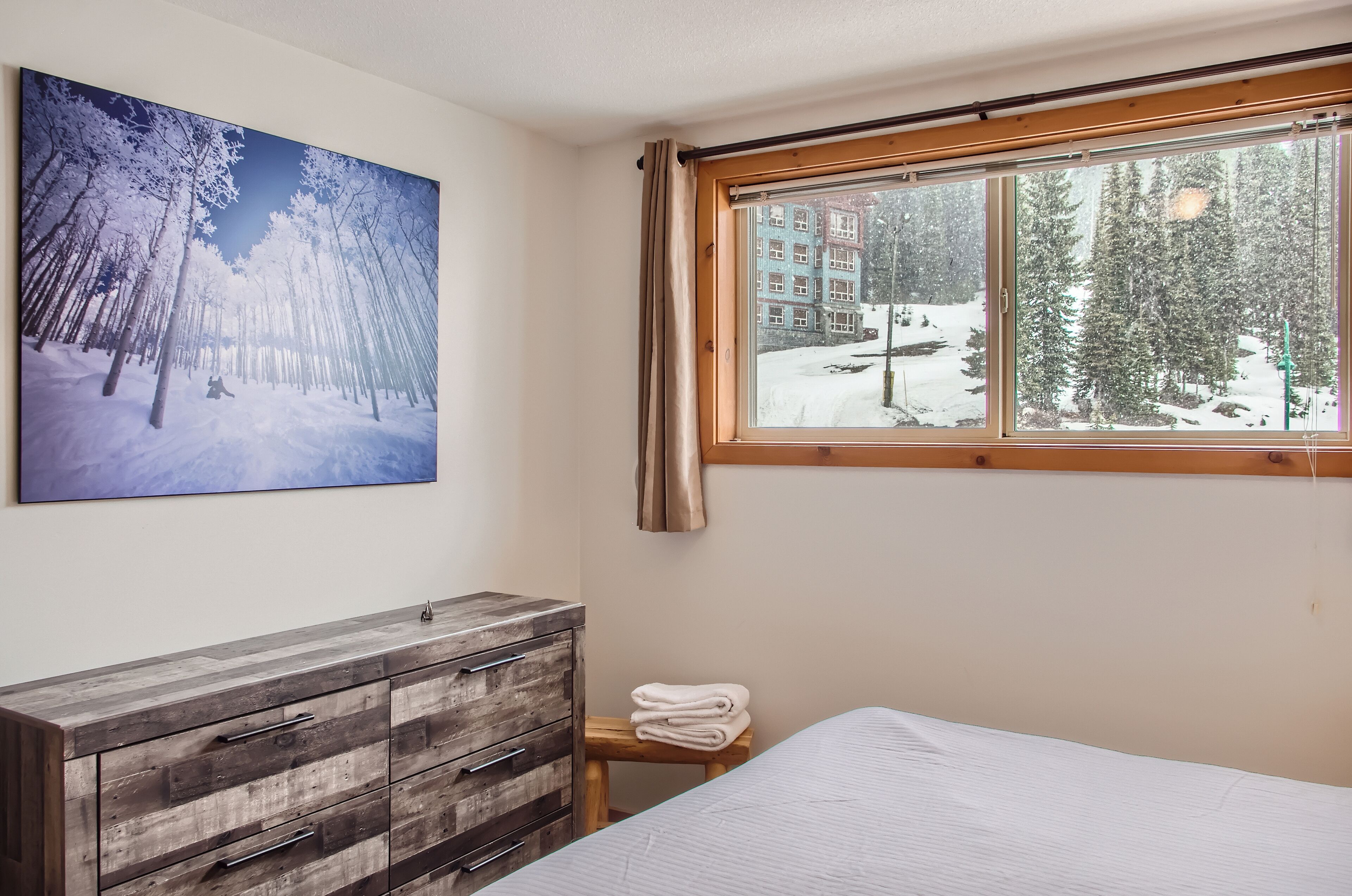 Une chambre avec une grande fenêtre, une commode en bois et un tableau représentant une forêt enneigée.