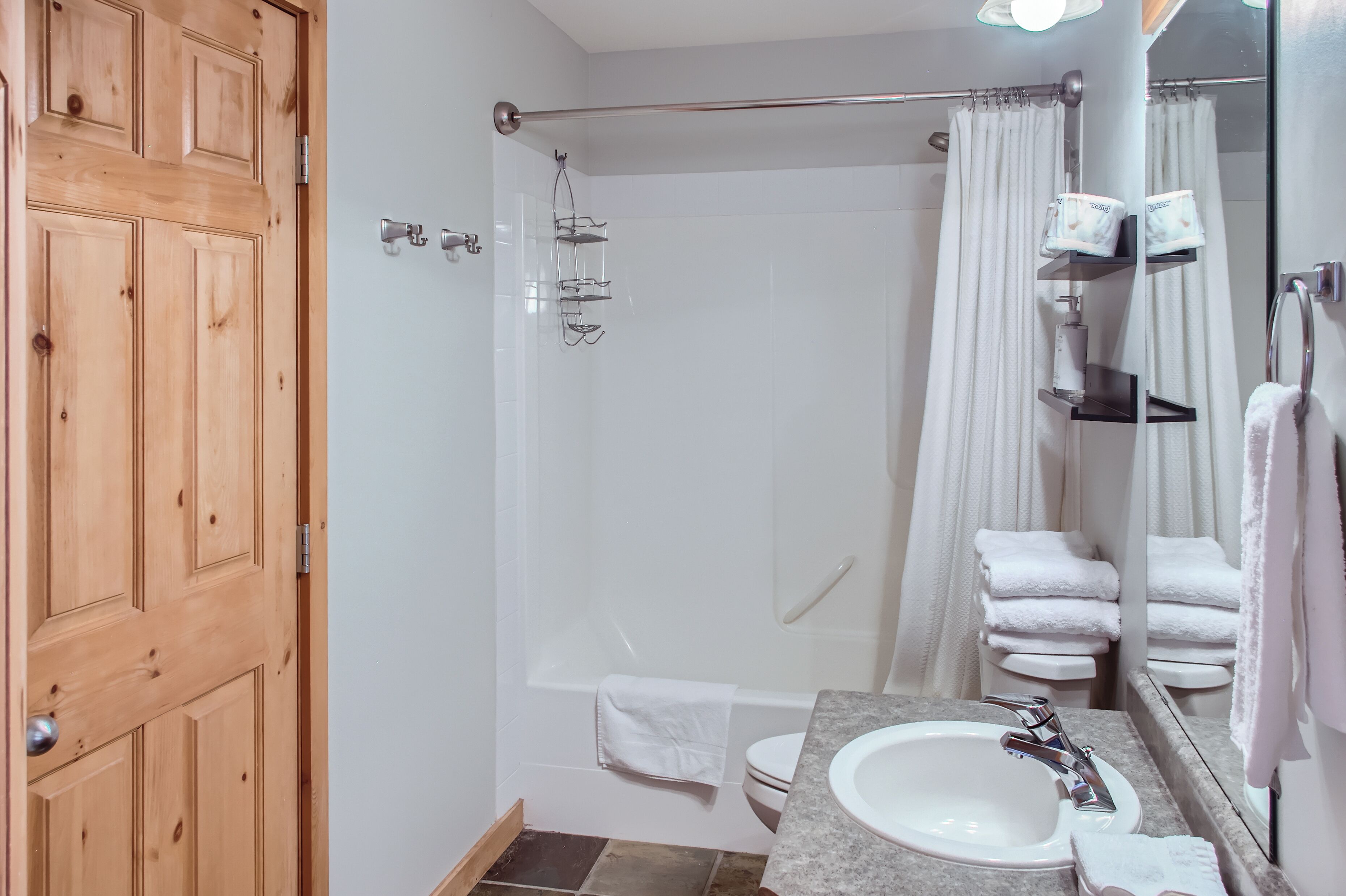 Ein Badezimmer mit Holztür, einer Duschkabine mit Glastür, einer Badewanne, einem Waschbecken mit Spiegel und einem Handtuchhalter mit gefalteten Handtüchern.