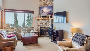 A cozy living room with a stone fireplace, a large window with a view of trees, and a mounted TV. - Snowy Creek by Okanagan Premier (Beaverdell)