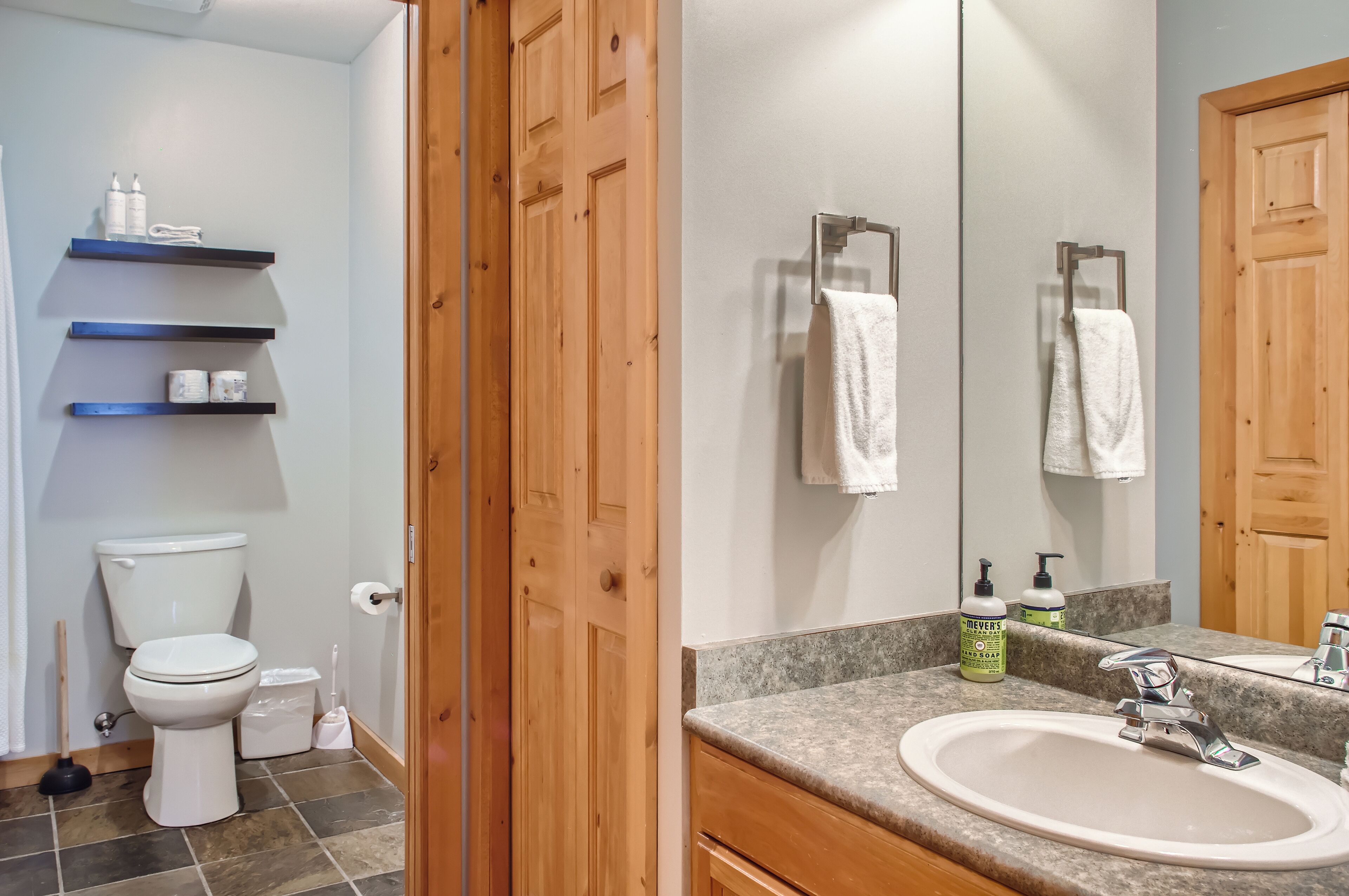 A bathroom with a white toilet, a sink with a mirror, and wooden shelves with toiletries.