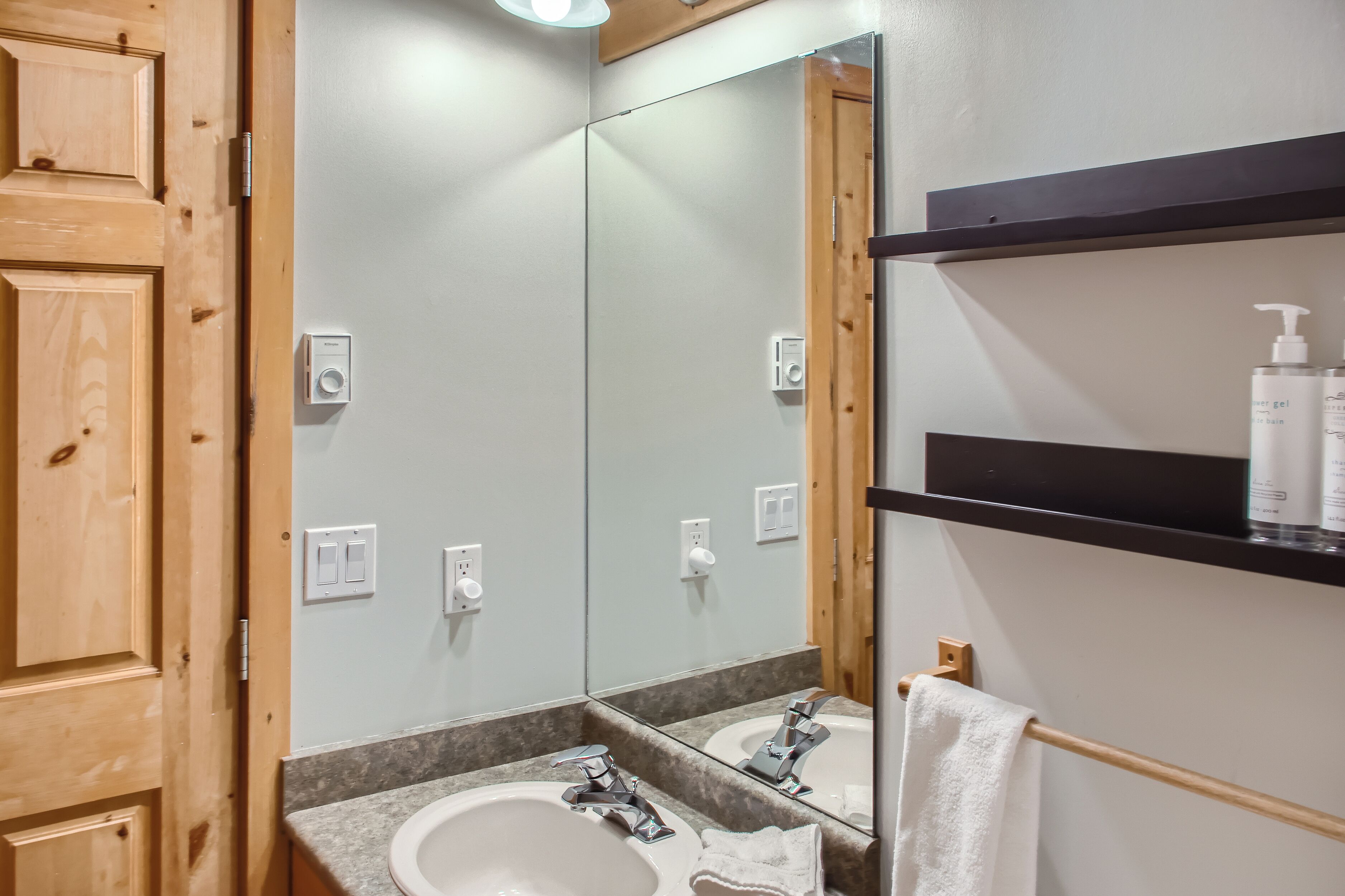 Une salle de bain avec une porte en bois, un lavabo blanc, un miroir et une étagère contenant des articles de toilette.
