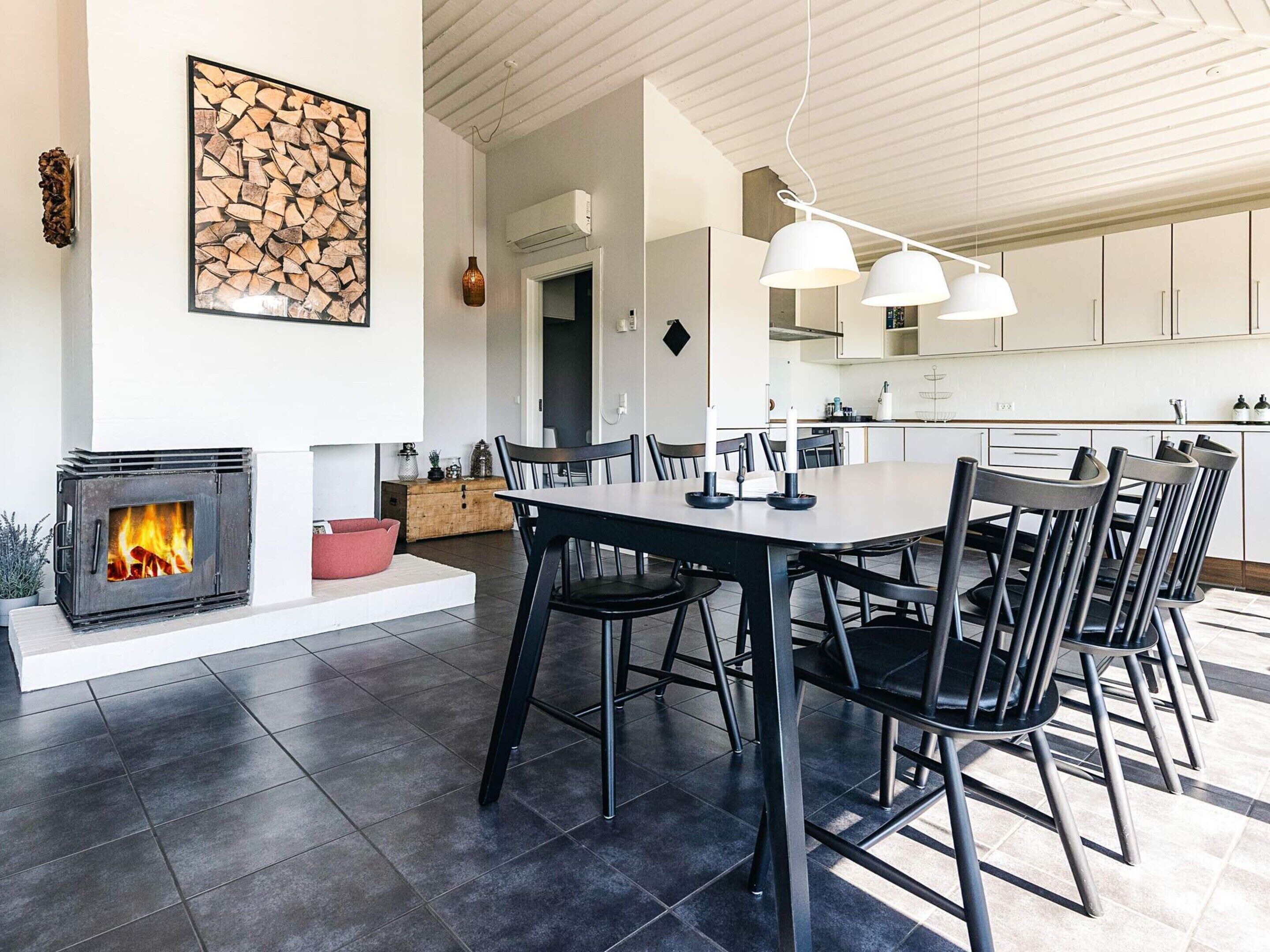 A modern kitchen with white cabinets, a dining area with a large table and chairs, and a fireplace with a visible fire.