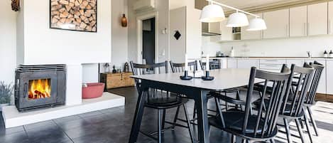 A modern kitchen with white cabinets, a dining area with a large table and chairs, and a fireplace with a visible fire.