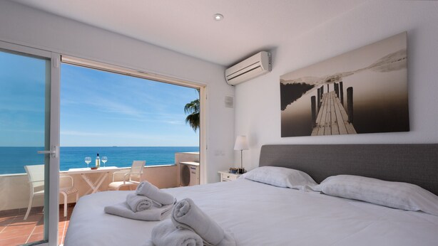 Une chambre avec un grand lit, une vue sur la mer, un coin repas avec des chaises et un tableau accroch é au mur.