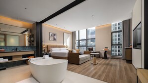 A modern hotel room with a large bathtub, a bed, a sofa, a small table, and a large window offering a city view.
