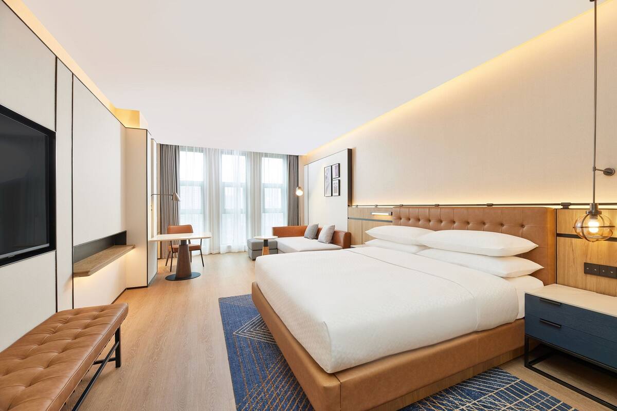 A modern hotel room with a large bed, a desk, a bench, and a TV.