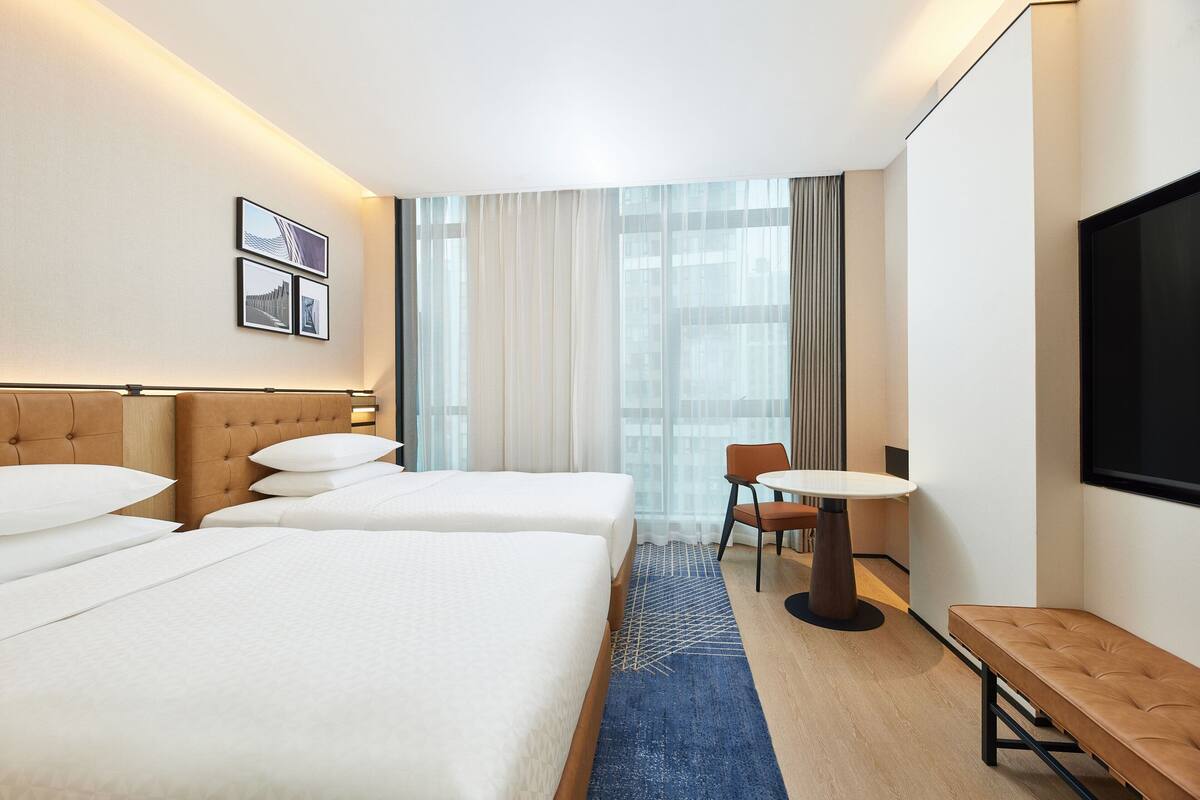A hotel room with two beds, a small table, a bench, a TV, and a window with curtains.