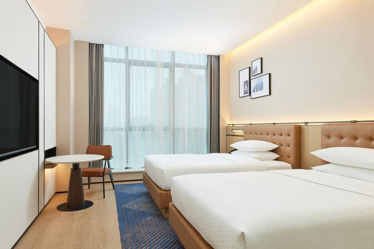 A hotel room with two beds, a TV, a small table, and a window with curtains.