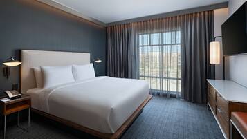 A hotel room with a large bed, a desk, a chair, a lamp, and a view of the outdoors through the window.