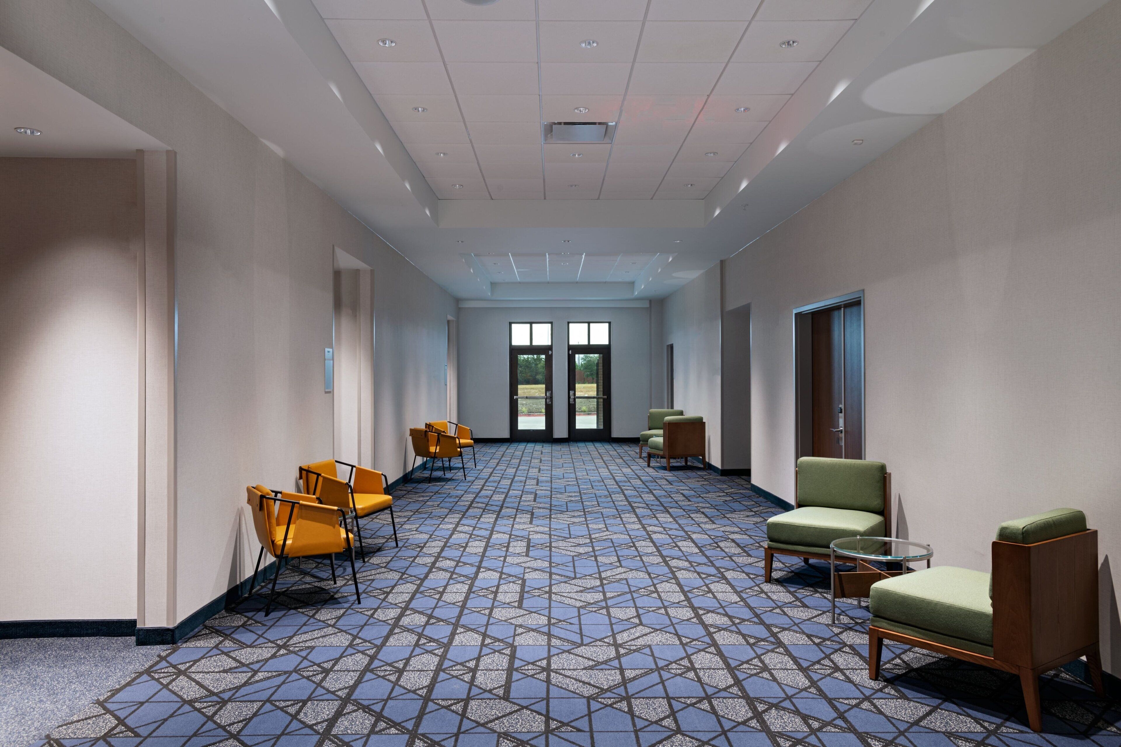 A hotel corridor with a patterned carpet, seating areas with chairs, and a view through the doors.