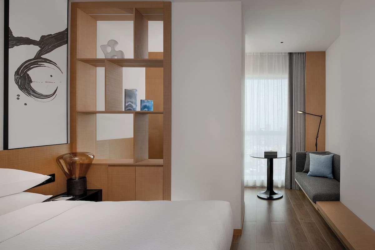 Moderni hotellihuone, jossa on sänky, puinen hyllykkö, pieni pöytä lampun kanssa ja tyynyillä varustettu sohva.
