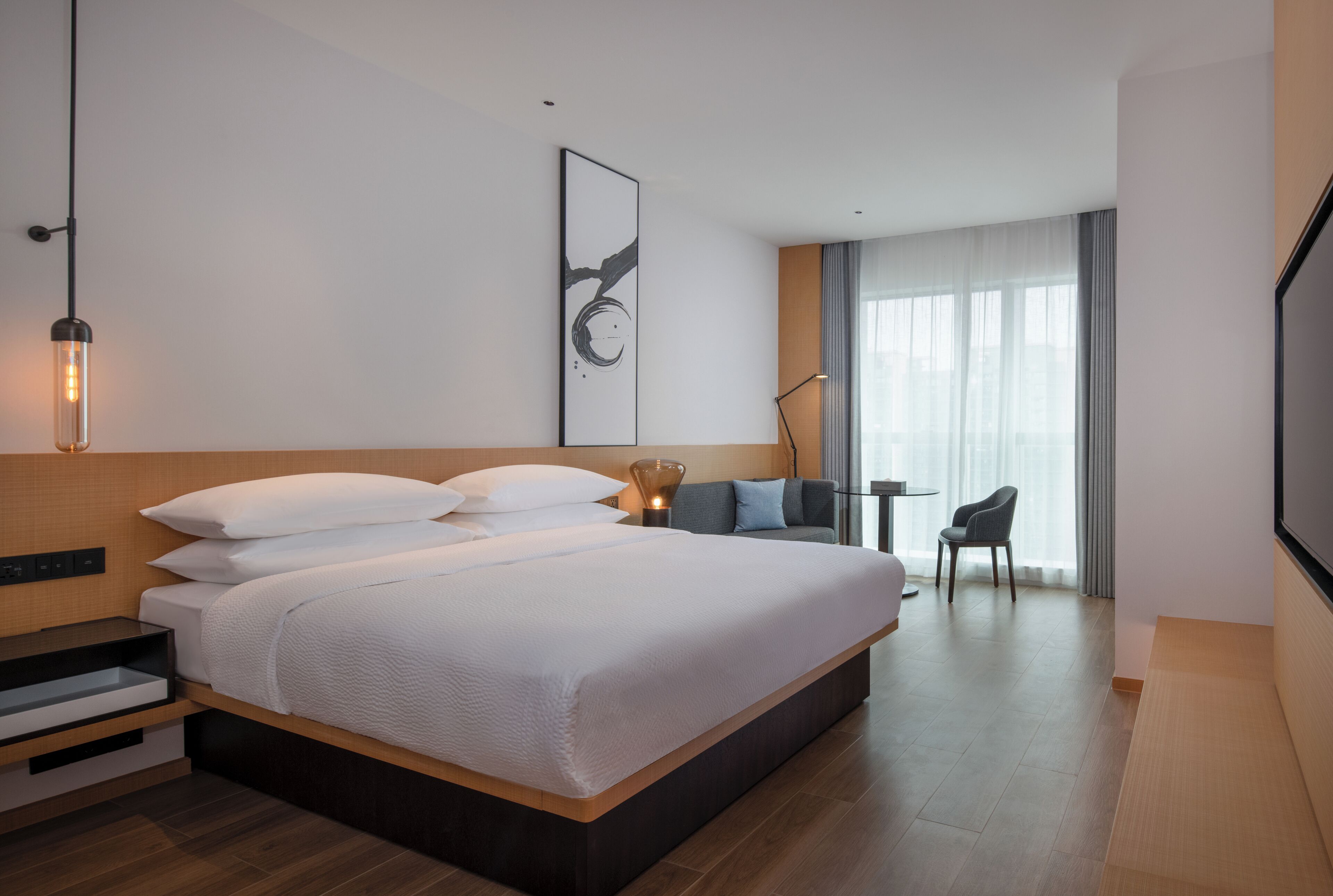 Ein modernes Hotelzimmer mit einem großen Bett, einem Schreibtisch, einem Sessel und einem großen Fenster mit Vorhängen.