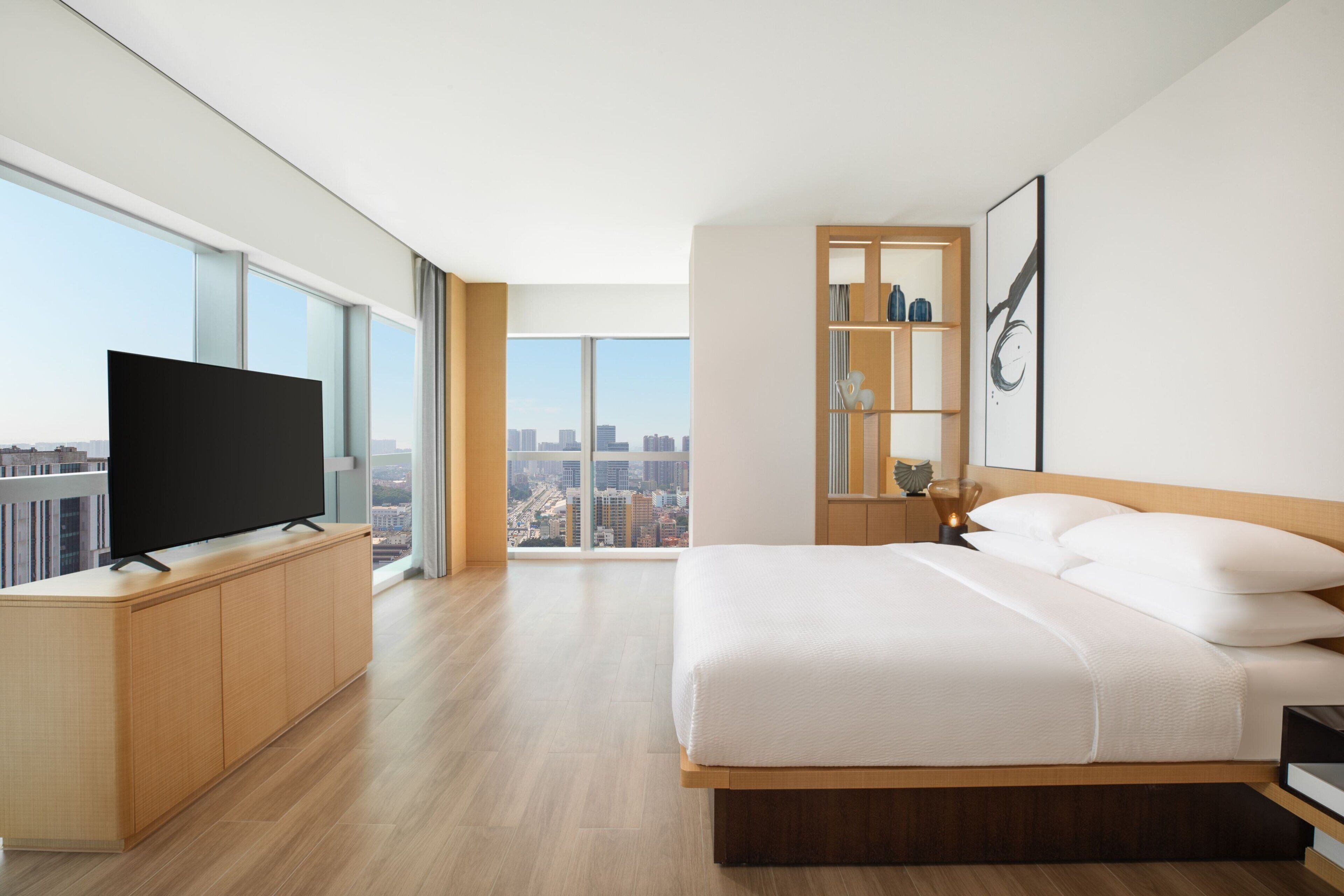 A modern hotel room with a large bed, a flat-screen TV on a wooden cabinet, a nightstand with a lamp, and a city view through large windows.