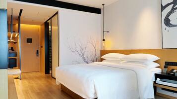 Habitación de hotel moderna con una cama grande, cabecera de madera, mesita de noche con lámpara y vistas al pasillo.