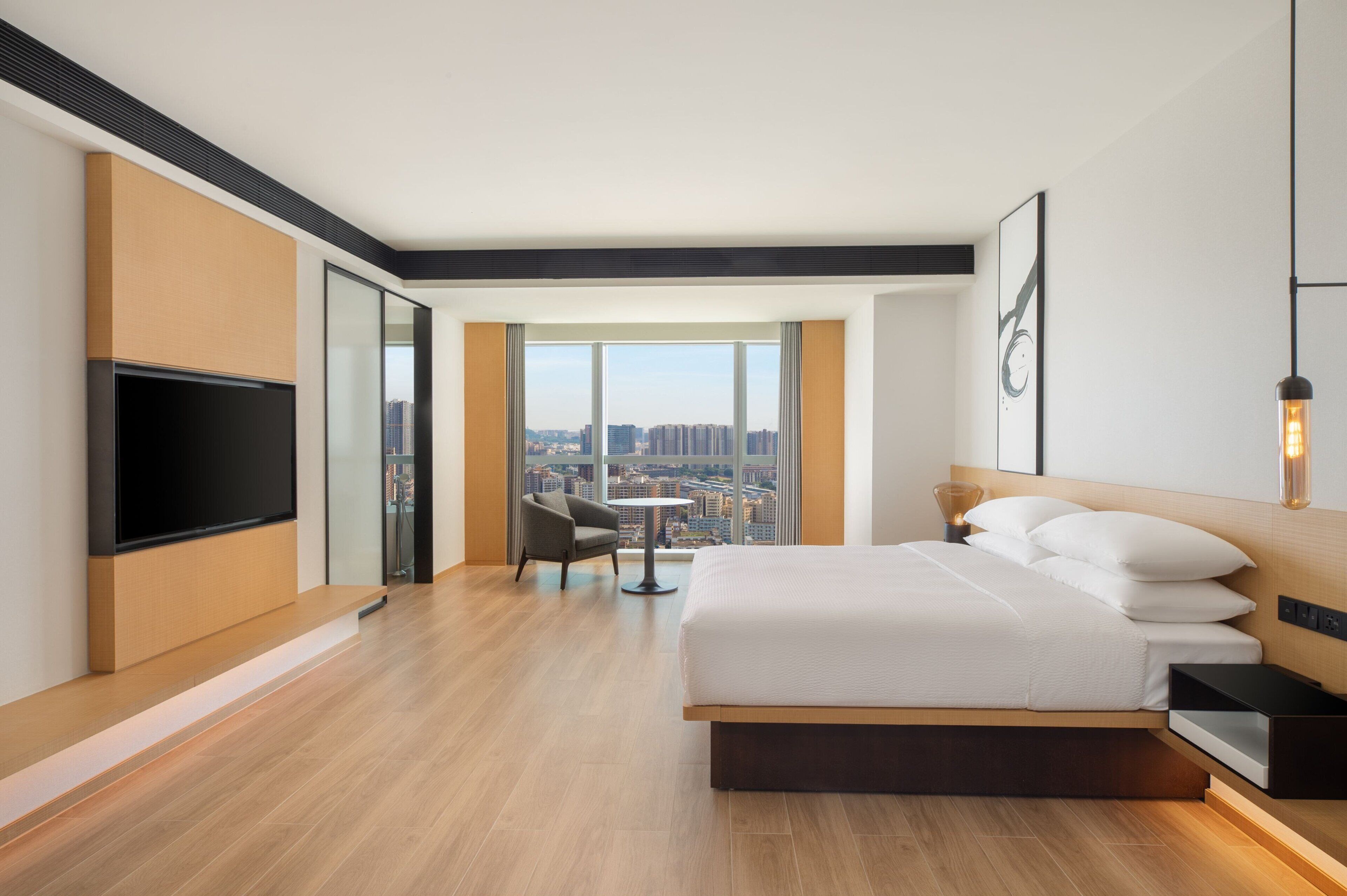 Una habitación de hotel moderna con una cama grande, un televisor de pantalla plana, vistas a la ciudad y una pequeña mesa con una silla.
