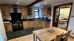 A modern kitchen with wooden cabinets, a black stove, and a dining area with a wooden table and chairs. - Lovely 3-bed House in Holmfirth (Holmfirth)
