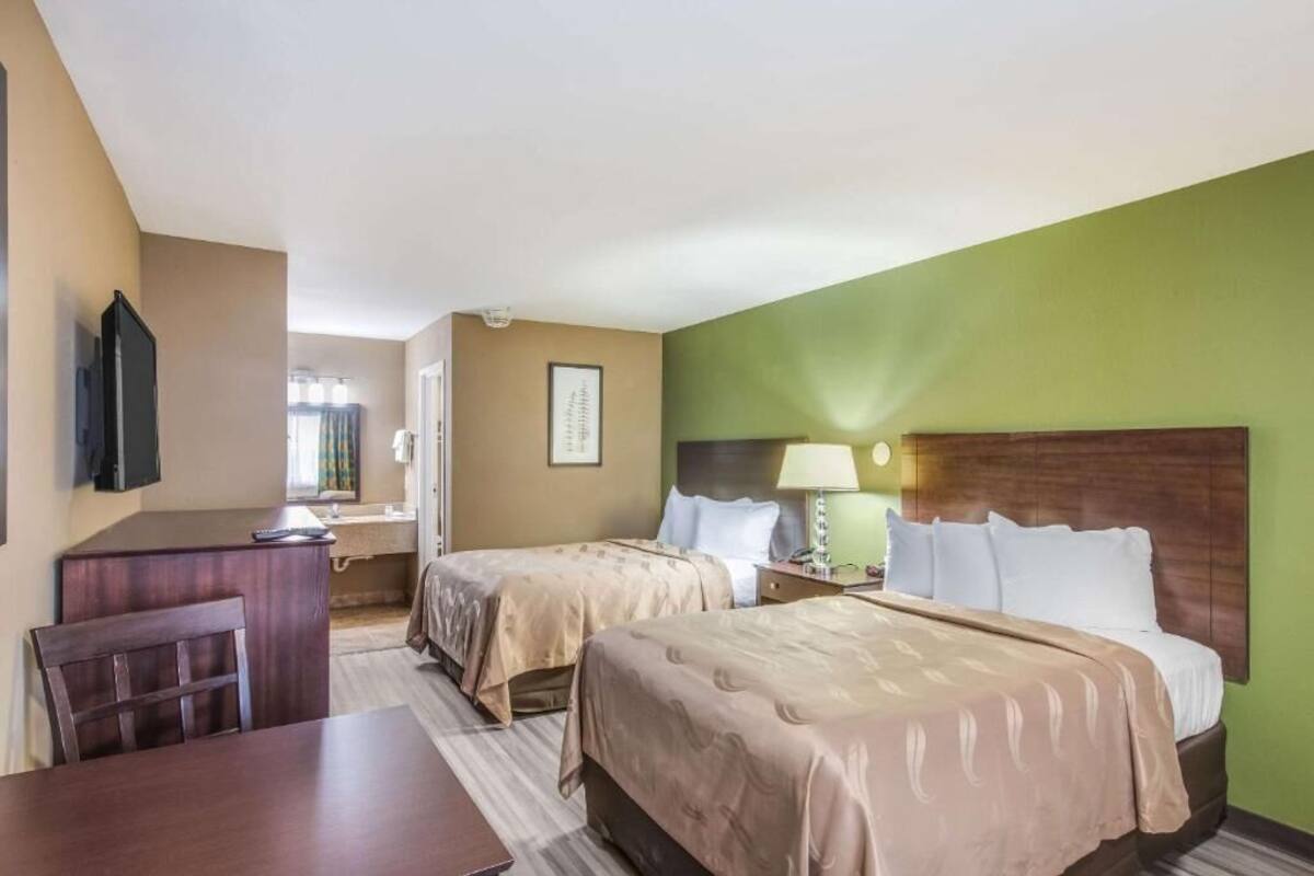 A hotel room with two beds, a desk, a chair, a TV, and a bathroom visible through an open door.