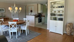 Eine geräumige Küche mit weißen Schränken, einem Essbereich mit Holztisch und weißen Stühlen sowie einer integrierten Regaleinheit mit Geschirr.