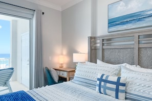 Une chambre d’hôtel avec un lit, une table de chevet, une chaise, une lampe et un tableau représentant l’océan.