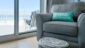 Ein graues Sofa mit einem türkisfarbenen Kissen, ein runder Hocker und ein großes Fenster mit Blick auf den Strand und Stühlen draußen.