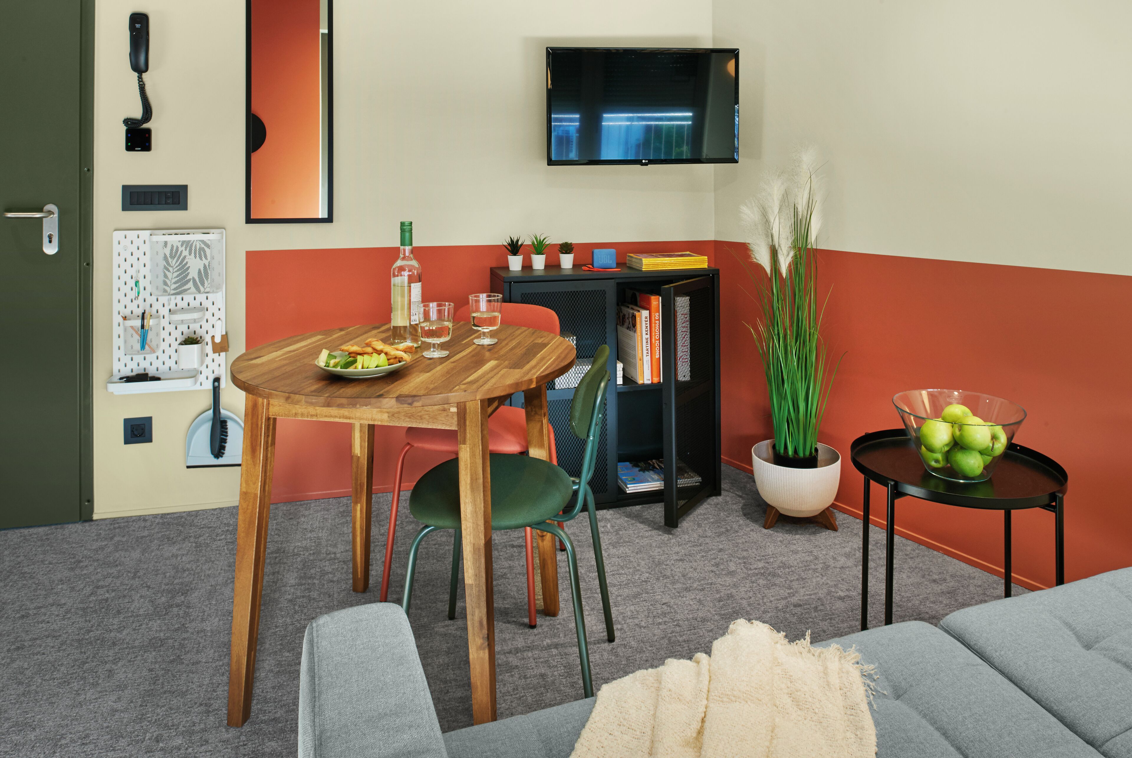 A room with a wooden dining table, green chairs, a sofa, a TV mounted on the wall, a bookshelf, and a small potted plant.