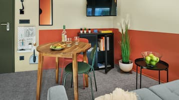 A room with a wooden dining table, green chairs, a sofa, a TV mounted on the wall, a bookshelf, and a small potted plant.