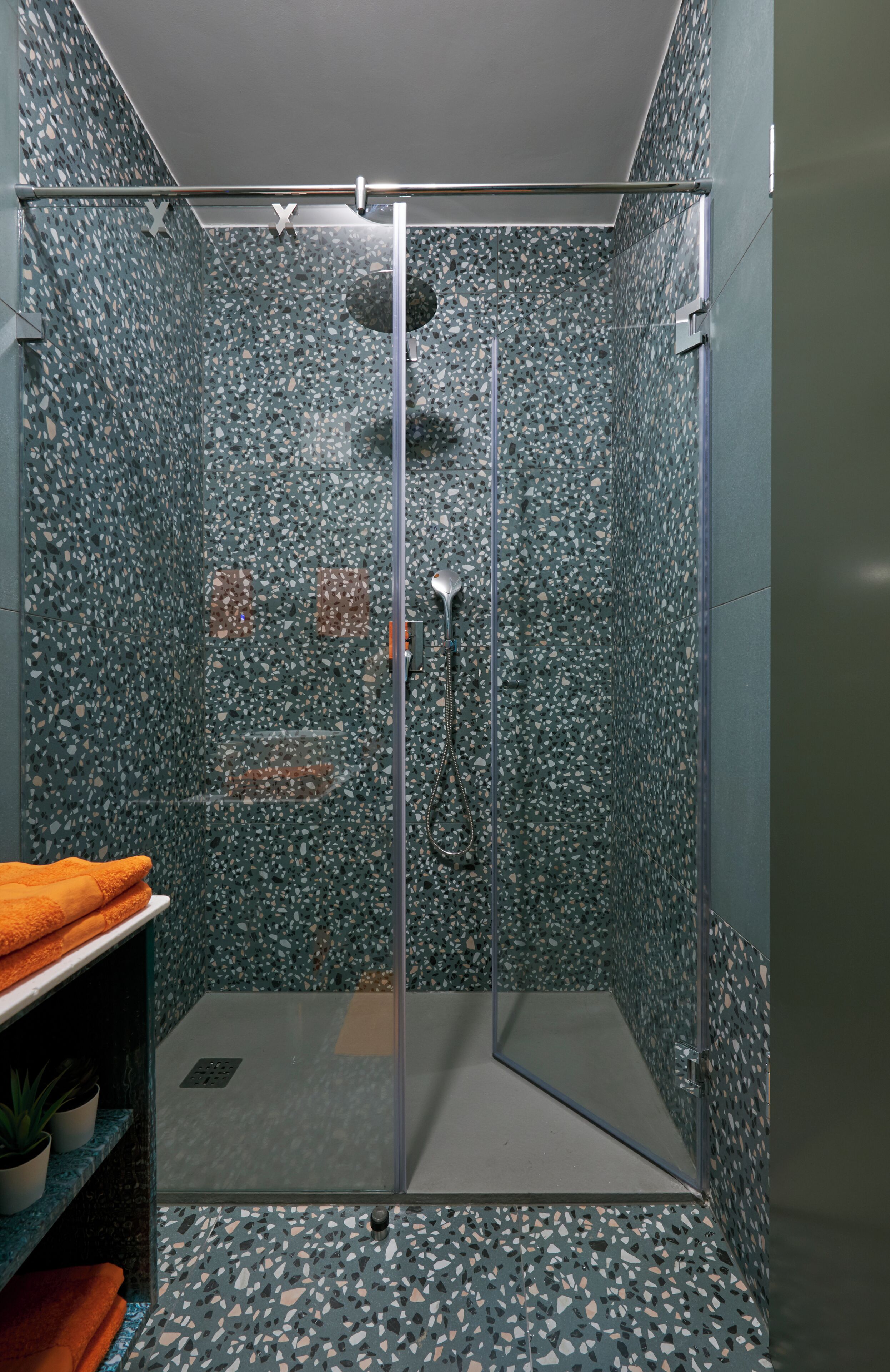 A glass-enclosed shower with a speckled tile design, a metal showerhead, and a shelf with towels.