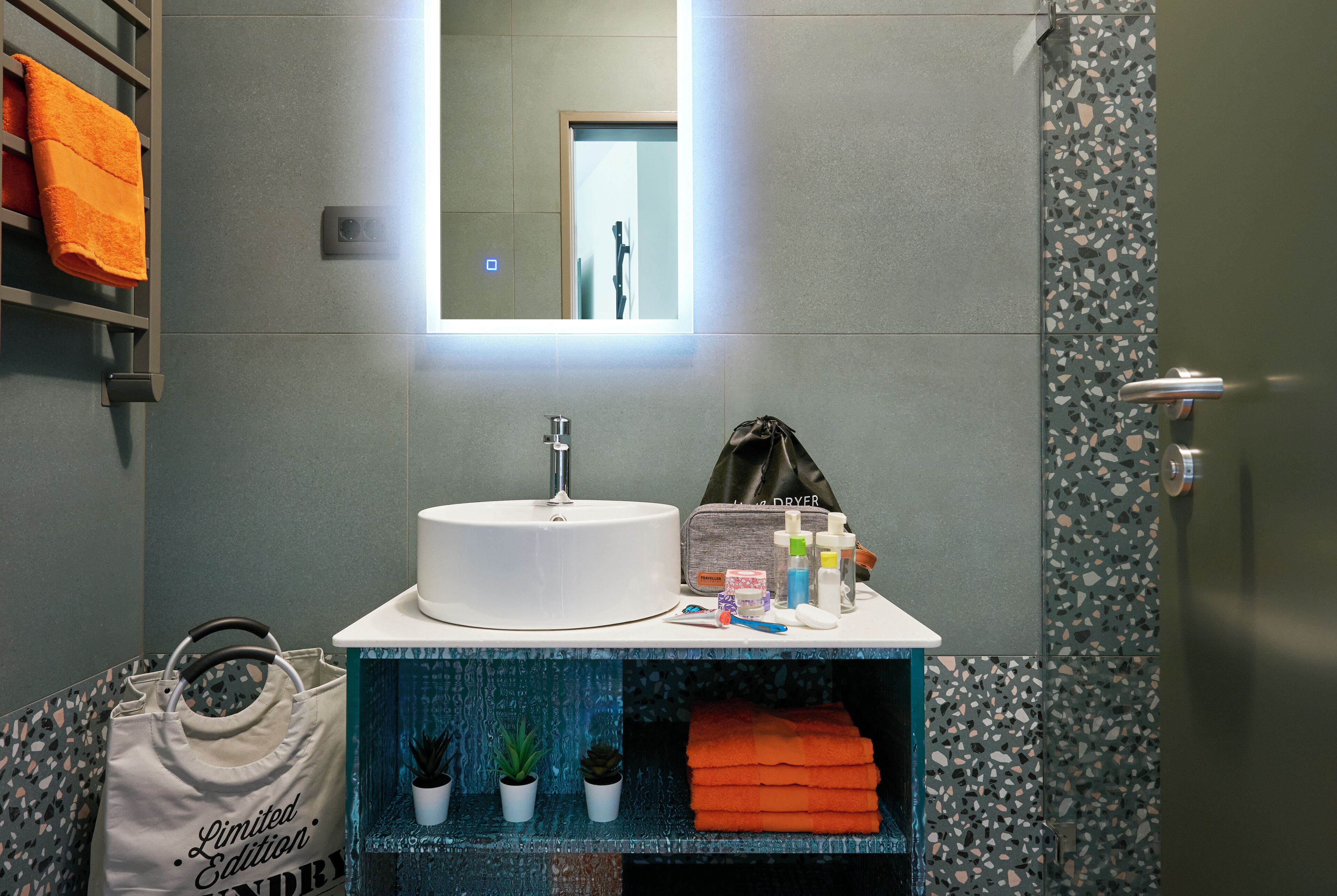 Ein Badezimmer mit einem weißen Waschbecken, einem Spiegel, einem Handtuchhalter mit einem orangefarbenen Handtuch, einer Tasche und einem Regal mit Pflanzen und gefalteten Handtüchern.
