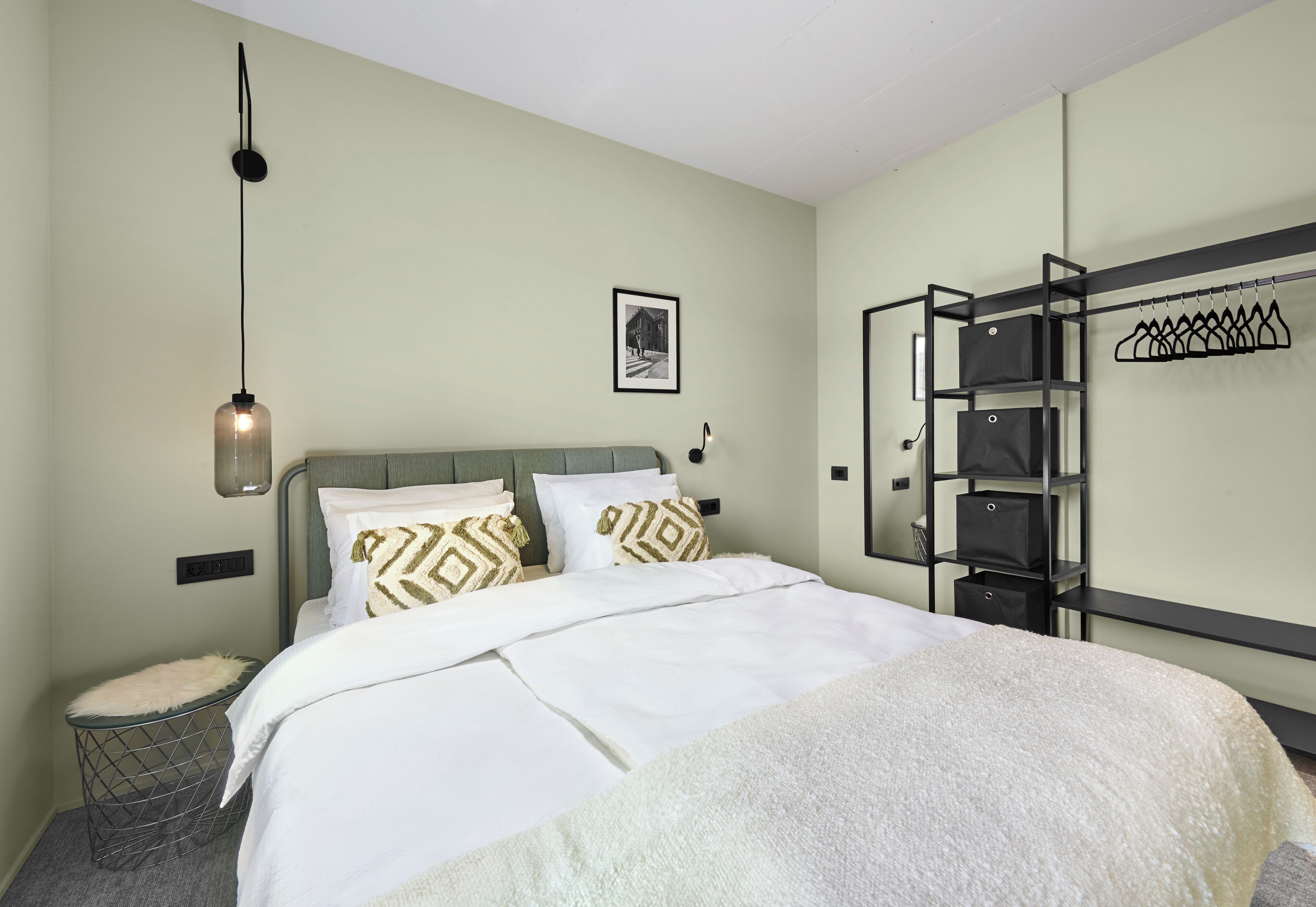 Ein ordentlich eingerichtetes Hotelzimmer mit Bett, einer Pendelleuchte, einem gerahmten Bild an der Wand und einem eingebauten Kleiderschrank mit Kleiderstange.