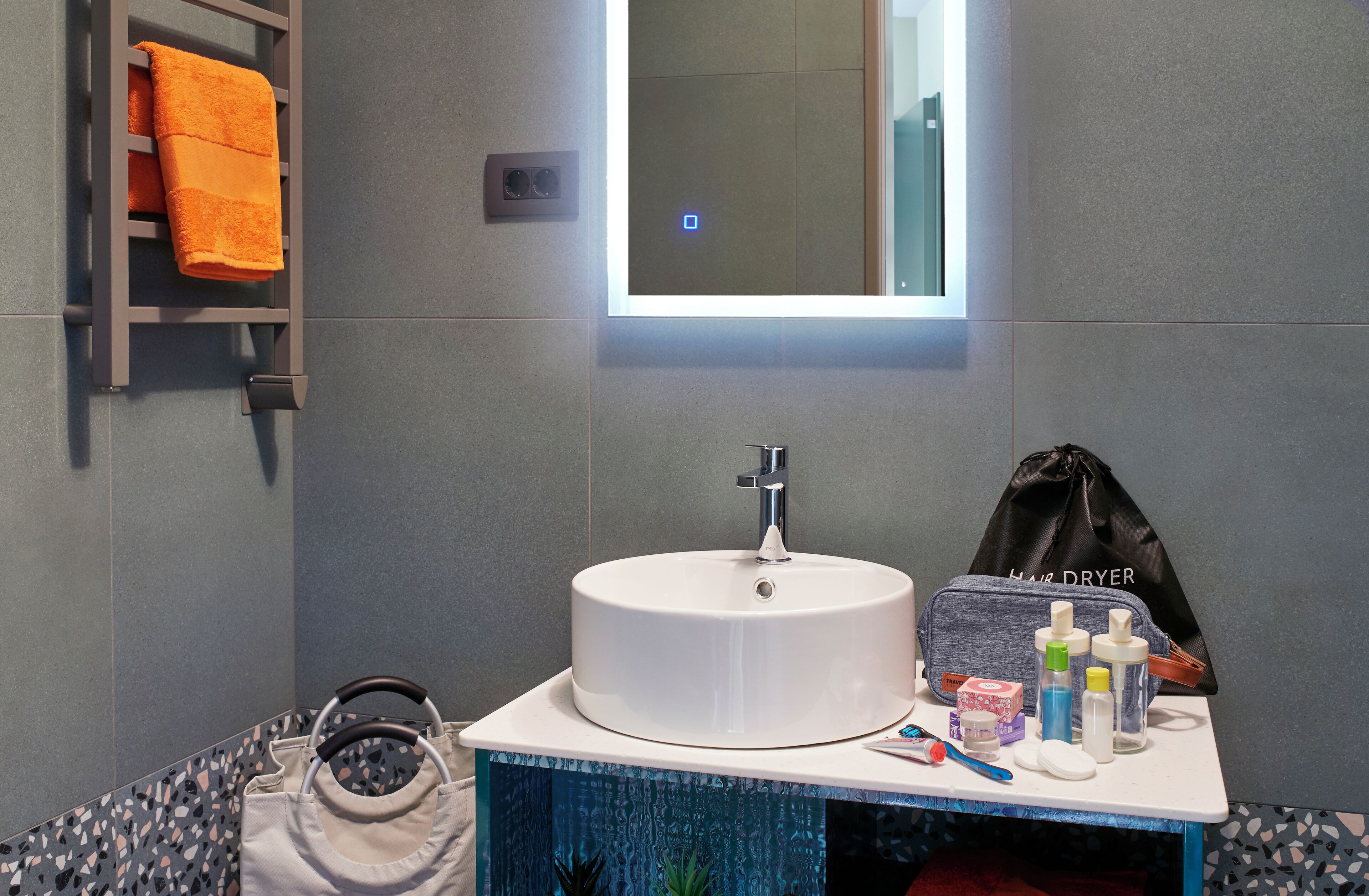 A bathroom with a round white sink, a towel rack with an orange towel, a hair dryer bag, and toiletries on the counter.