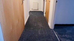 Ein schmaler Korridor mit Teppichböden, Holztüren und einem Heizkörper an der Wand.